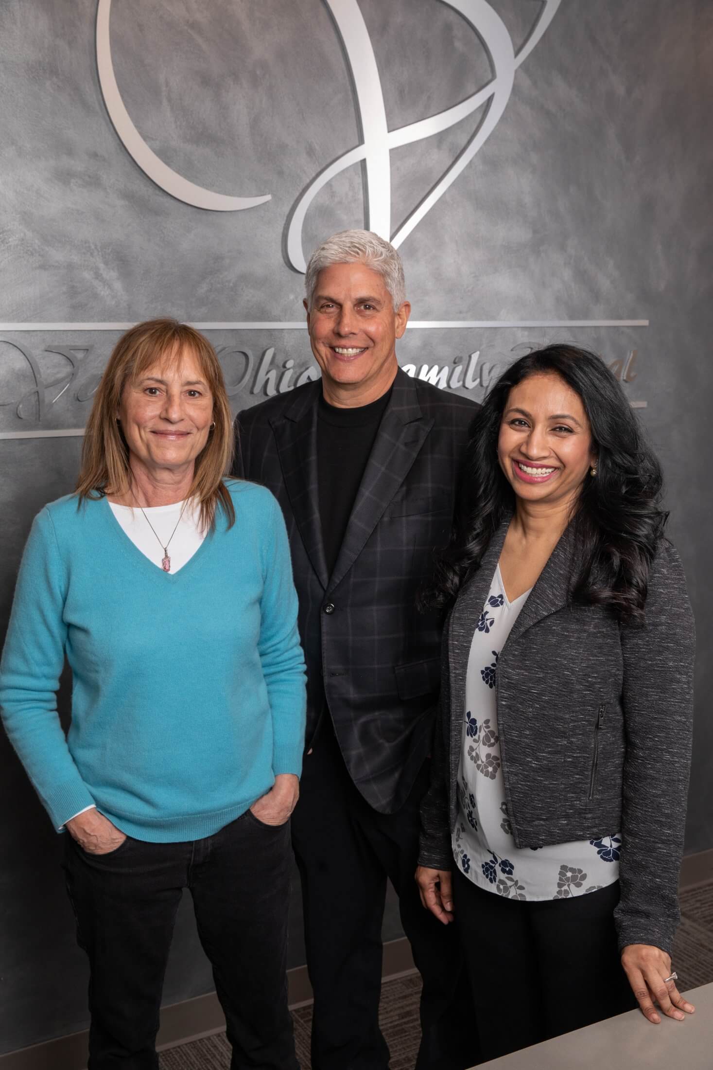Jane, Dr. Vadhi, and Dr. Urell standing next to the office wall with the practice logo behind them